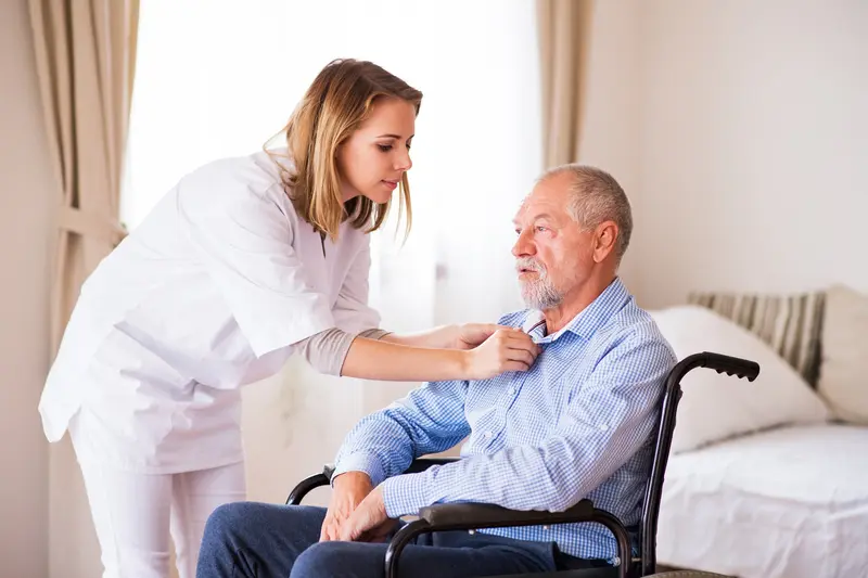 A caregiver helping an older man in a wheelchair with his shirt collar