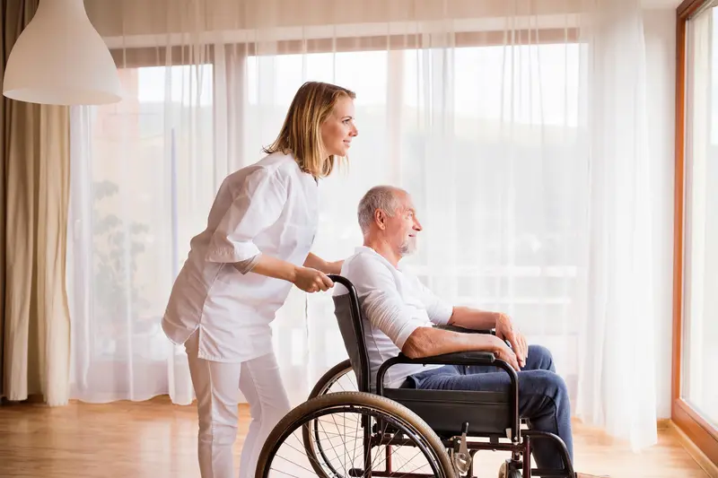 A caregiver pushing an older man in a wheelchair beside a sunny window