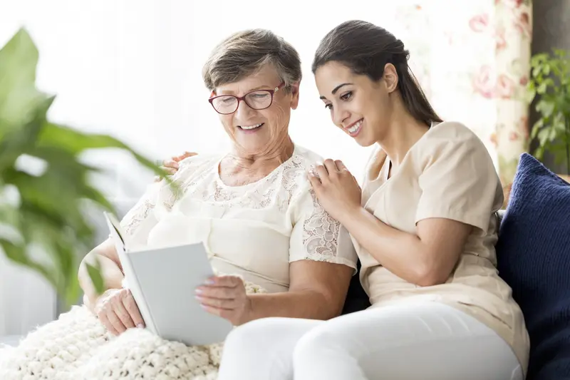 A smiling caregiver reading a book with an older woman on her sofa