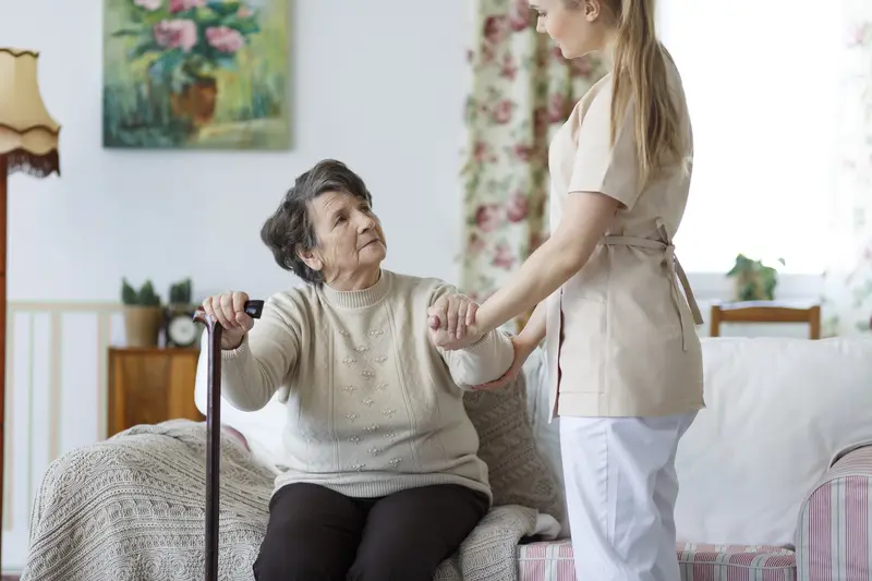 A caregiver helping an older woman with a walking stick rise from her armchair
