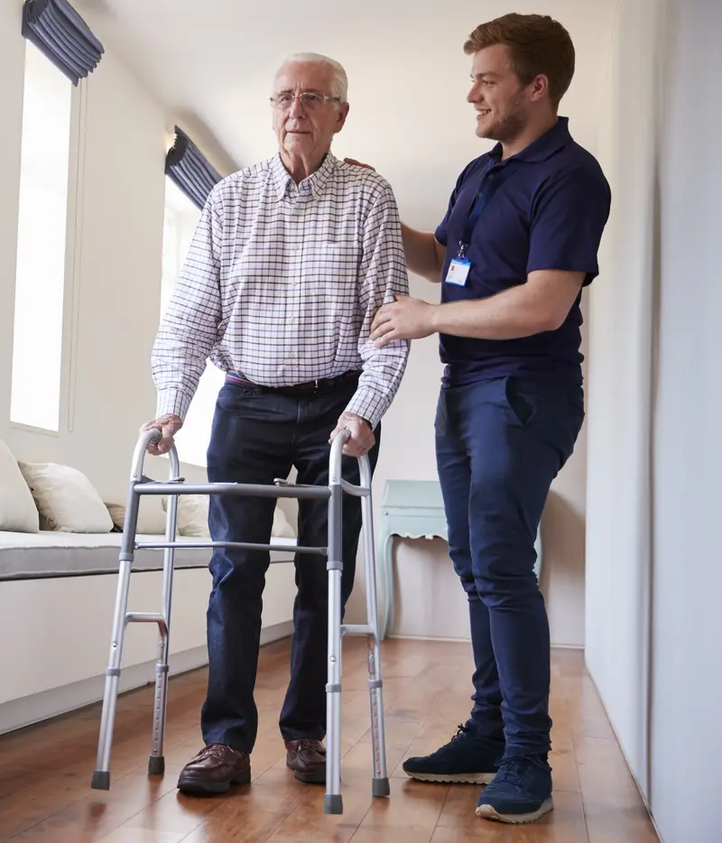 A young male caregiver supporting an older man walking with a frame