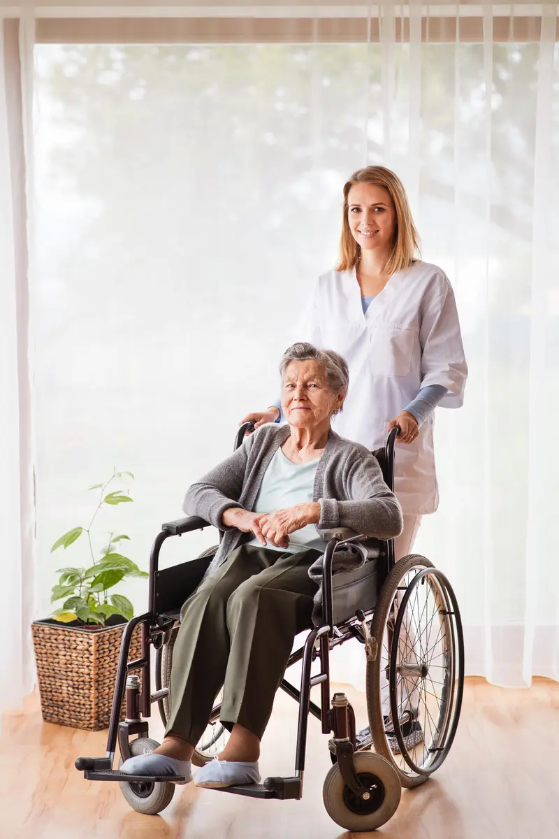 A caregiver standing beside a smiling older woman in a wheelchair by a sunny window